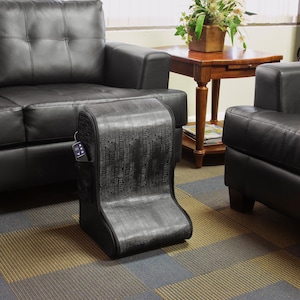 May include: A black foot massager with a textured fabric cover. The massager has a built-in remote control and is positioned in front of a black leather sofa.