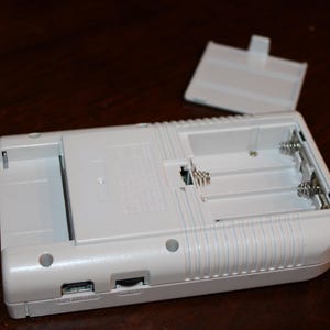 May include: A white handheld gaming device with an open battery compartment and detached cover. The device has a rectangular shape with rounded edges and a series of vertical lines on the side. The battery compartment is designed to hold multiple batteries.
