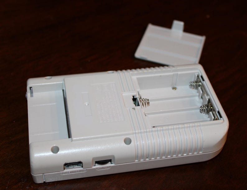 May include: A white handheld gaming device with an open battery compartment and detached cover. The device has a rectangular shape with rounded edges and a series of vertical lines on the side. The battery compartment is designed to hold multiple batteries.
