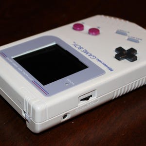 May include: A white Nintendo Game Boy handheld video game console. The device features a black screen, purple buttons, and a directional pad. The text "Nintendo GAME BOY" is printed on the top. The console is a classic retro gaming device.