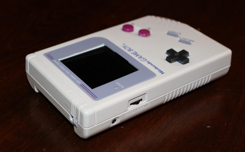 May include: A white Nintendo Game Boy handheld video game console. The device features a black screen, purple buttons, and a directional pad. The text "Nintendo GAME BOY" is printed on the top. The console is a classic retro gaming device.