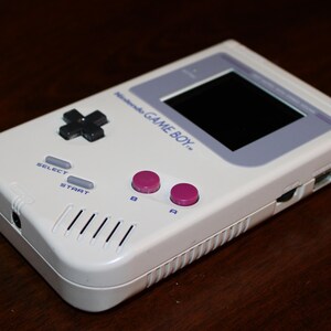 May include: A white Nintendo Game Boy handheld console. The device features a black directional pad, gray select and start buttons, and pink A and B buttons. The screen is black, and the text "Nintendo GAME BOY" is visible.