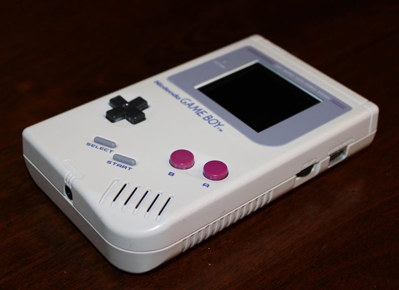 May include: A white Nintendo Game Boy handheld console. The device features a black directional pad, gray select and start buttons, and pink A and B buttons. The screen is black, and the text "Nintendo GAME BOY" is visible.