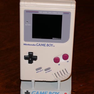 May include: A cream-colored Nintendo Game Boy handheld console, with a black screen, gray accents, and pink buttons. The console is on a matching stand with the words "GAME BOY" printed on it. The console has the text "DOT MATRIX WITH STEREO SOUND".