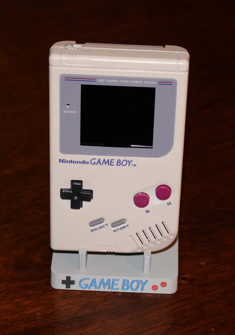 May include: A cream-colored Nintendo Game Boy handheld console, with a black screen, gray accents, and pink buttons. The console is on a matching stand with the words "GAME BOY" printed on it. The console has the text "DOT MATRIX WITH STEREO SOUND".
