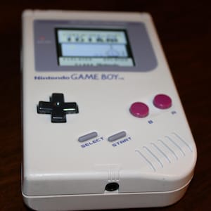 May include: A white Nintendo Game Boy handheld console. The device features a black directional pad, pink A and B buttons, and gray select and start buttons. The screen displays a game. The text "Nintendo GAME BOY" is visible.