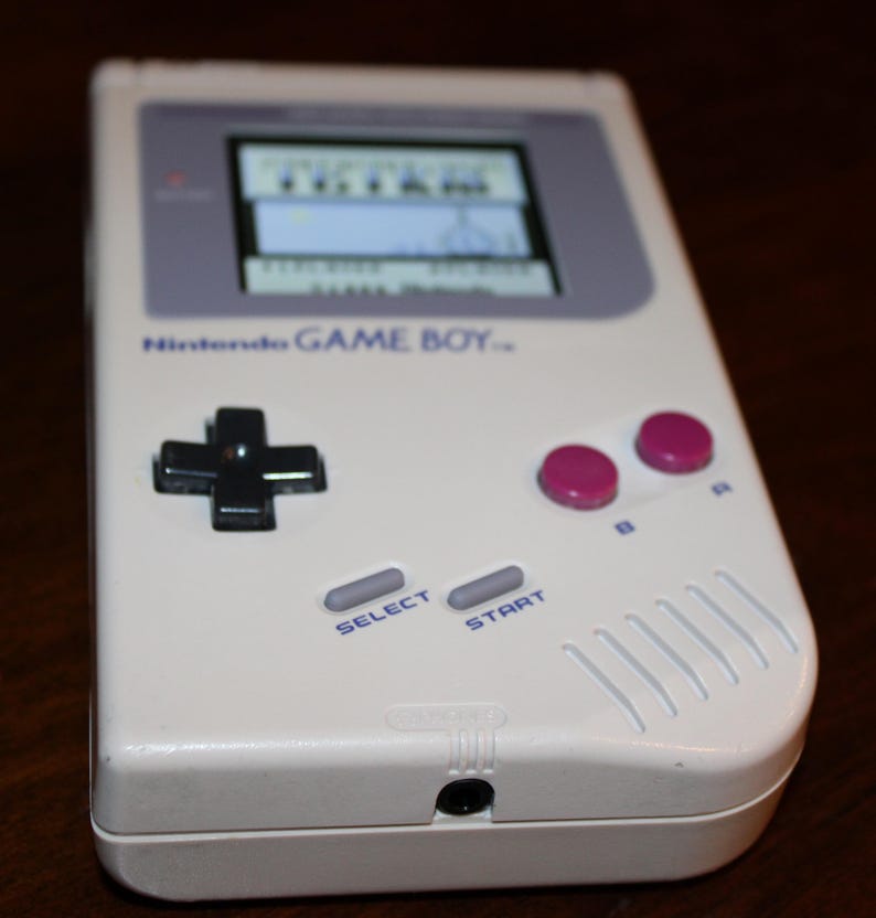 May include: A white Nintendo Game Boy handheld console. The device features a black directional pad, pink A and B buttons, and gray select and start buttons. The screen displays a game. The text "Nintendo GAME BOY" is visible.