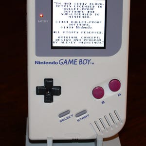 May include: A classic off-white Nintendo Game Boy handheld console, displayed on a stand. The screen displays text, including "Tetris" and "Nintendo." Features include a directional pad, buttons, and the text "GAME BOY" on the stand.