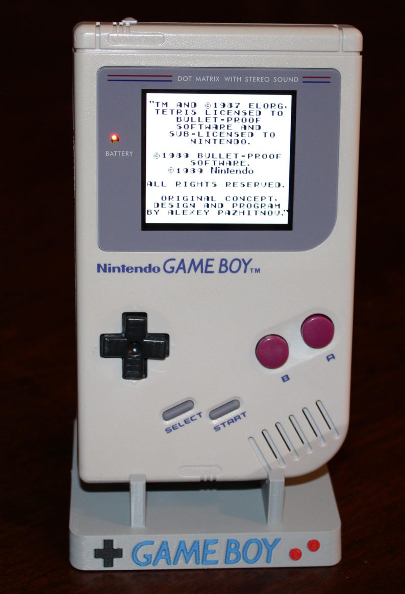 May include: A classic off-white Nintendo Game Boy handheld console, displayed on a stand. The screen displays text, including "Tetris" and "Nintendo." Features include a directional pad, buttons, and the text "GAME BOY" on the stand.