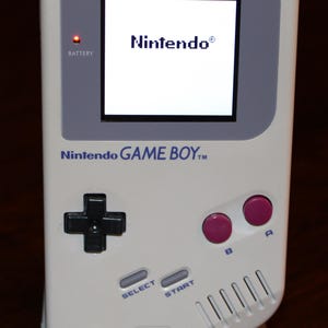 May include: A white Nintendo Game Boy handheld console, featuring a black directional pad, pink A and B buttons, and a screen displaying the Nintendo logo. The console has the text "Nintendo GAME BOY" and "DOT MATRIX WITH STEREO SOUND".