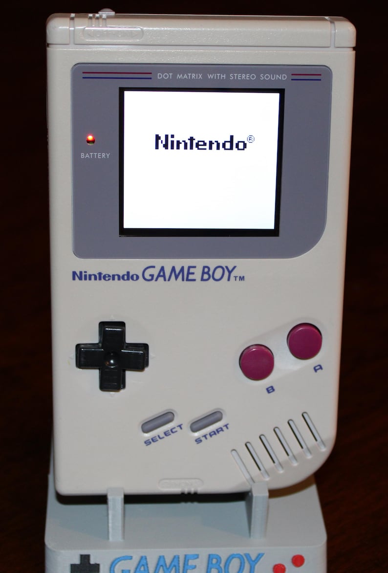 May include: A white Nintendo Game Boy handheld console, featuring a black directional pad, pink A and B buttons, and a screen displaying the Nintendo logo. The console has the text "Nintendo GAME BOY" and "DOT MATRIX WITH STEREO SOUND".