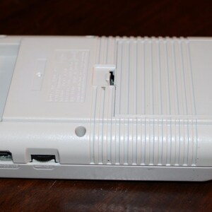 May include: A white handheld gaming console, viewed from the back, with a rectangular shape and rounded corners. The device has a cartridge slot, a power port, and a volume slider. The back features a battery compartment and a ridged design.