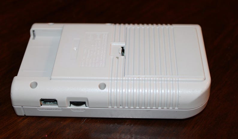 May include: A white handheld gaming console, viewed from the back, with a rectangular shape and rounded corners. The device has a cartridge slot, a power port, and a volume slider. The back features a battery compartment and a ridged design.