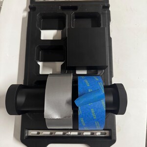 Puede incluir: Organizador de herramientas negro Milwaukee Packout con un logotipo rojo y blanco. El organizador contiene un rollo de cinta adhesiva plateada y un rollo de cinta de pintor azul. Diseñado para guardar herramientas y suministros.