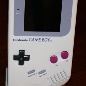May include: A white Nintendo Game Boy handheld console, featuring a black directional pad, pink A and B buttons, and a screen with the text "DOT MATRIX WITH STEREO SOUND." The console is on a white stand with the text "GAME BOY."