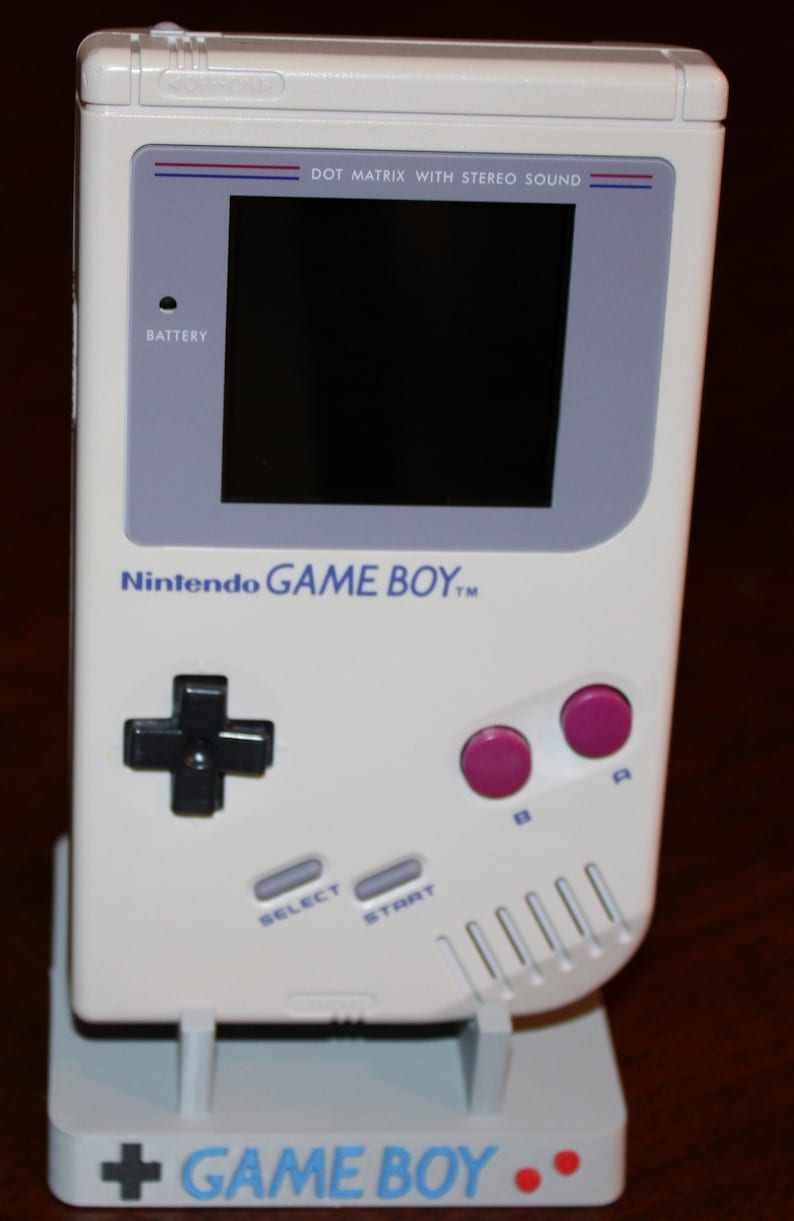 May include: A white Nintendo Game Boy handheld console, featuring a black directional pad, pink A and B buttons, and a screen with the text "DOT MATRIX WITH STEREO SOUND." The console is on a white stand with the text "GAME BOY."