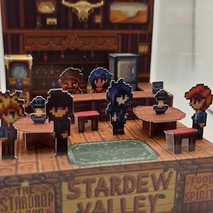 May include: A diorama of the Stardrop Saloon from Stardew Valley. The scene features pixelated characters, tables, and a bar. The saloon's name is displayed on a wooden sign. The interior is detailed with a bar, stools, and a green rug.