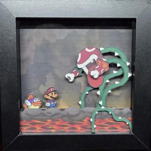 May include: Framed artwork featuring a scene from a classic video game. The artwork depicts a red and white Piranha Plant with green vines, facing Mario and a character with wings. The background is a cloudy sky and lava.