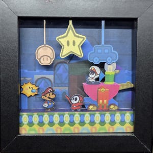 May include: A framed shadow box diorama featuring characters and elements from the Super Mario video game series. The scene includes Mario, a Shy Guy, a mushroom, a star, and a car. The background is a blue cityscape.