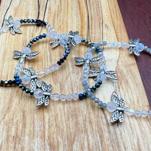 May include: Three beaded bracelets with dragonfly charms. The bracelets feature clear, black, and gray beads, with silver-colored dragonfly charms. The bracelets are displayed on a wooden surface.