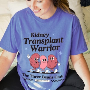 May include: A purple t-shirt featuring the text "Kidney Transplant Warrior" and a graphic of three cartoon kidney beans walking. The text "The Three Beans Club" is below the graphic, with the words "Saved by a Spare" underneath.