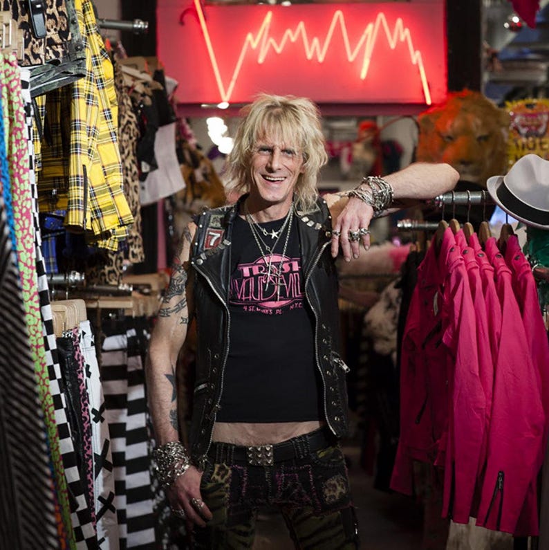 May include: A man with blonde hair and a black leather waistcoat is standing in a clothing store. He is wearing a black t-shirt with the words "True Vaudeville" printed on it. He is also wearing a black belt with a silver buckle and black trousers with a pink and green pattern. The store is filled with colourful clothing and accessories.