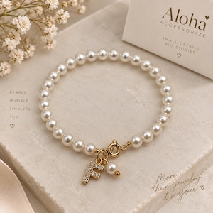 Puede incluir: Una pulsera de cuentas de perlas blancas con un cierre circular dorado y un dije colgante con la letra F dorada incrustada con pequeños cristales transparentes, junto a un pequeño dije de perla. El texto Aloha ACCESSORIZE es visible.