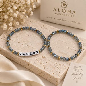Puede incluir: Dos pulseras de cuentas con cristales facetados de color azul grisáceo y pequeñas cuentas metálicas doradas. Una pulsera incluye cuentas cuadradas blancas con letras que forman VALERY. El nombre ALOHA ACCESSORIZE aparece en una caja.