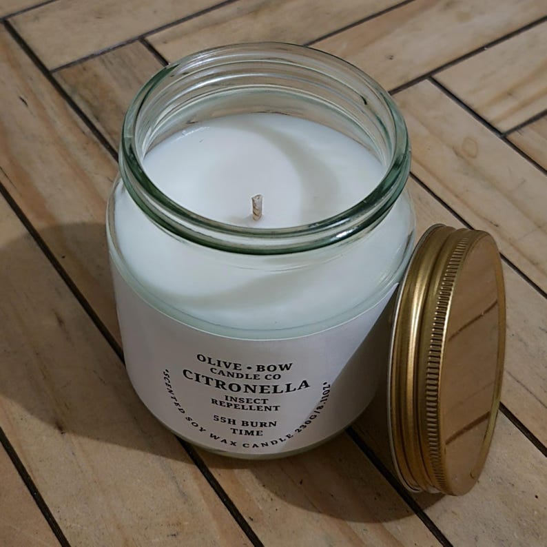 Peut inclure: Une bougie en pot de verre avec un int&eacute;rieur en cire blanche et un couvercle dor&eacute;. L'&eacute;tiquette indique "OLIVE + BOW CANDLE CO CITRONELLA INSECT REPELLENT". La bougie est une bougie parfum&eacute;e &agrave; la cire de soja avec 55 heures de combustion.