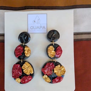 Puede incluir: Pendientes florales negros, rojos y dorados. Los pendientes tienen un círculo superior e inferior negro brillante, con un círculo central con diseños florales rojos y dorados. Se presentan en una tarjeta blanca con la marca "GUAPA".