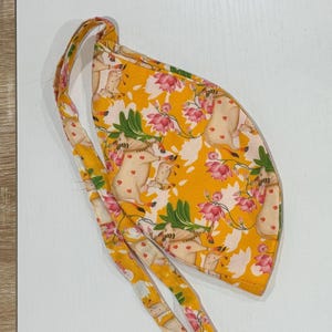 May include: A yellow patterned surgical scrub cap with a tie closure. The cap features a repeating pattern of light brown animals with heart-shaped markings, pink lotus flowers, and green leaves. The ties are a cream color with a similar pattern.