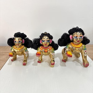 May include: Three golden figurines of a deity, each with black hair and detailed facial features. The figurines are in a crawling position, with painted details on their bodies and colorful accents on their jewelry. They are set against a neutral background.