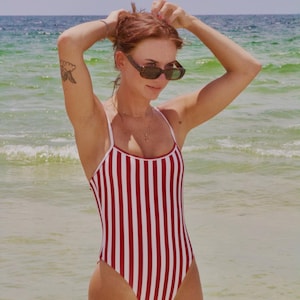 May include: A red and white striped swimsuit with thin white straps. The swimsuit is a one-piece design. The person is wearing sunglasses and standing in the sea.