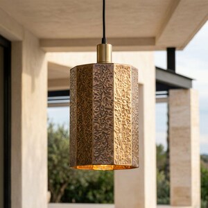 May include: A cylindrical pendant light fitting featuring a textured, hammered brass finish with a repeating organic pattern. The lamp is suspended by a black cord and includes a smooth, cylindrical brass cap at the top where the cord connects.