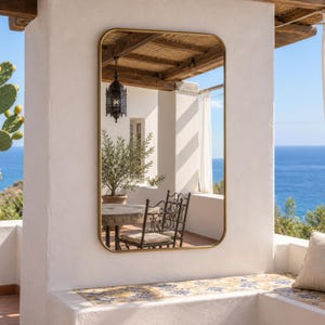May include: A rectangular wall mirror with rounded corners and a thin gold-toned metal frame. The mirror is mounted on a white pillar and reflects a patio area with a wooden ceiling, a hanging lantern, a small tree, and outdoor furniture.