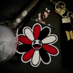 May include: A black, white, and red embroidered flower patch with a smiling face in the center. The patch is on a black surface.