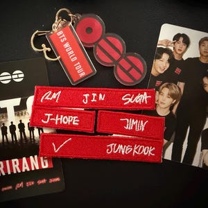 Könnte beinhalten: Eine Sammlung von BTS-Fanartikeln, bestehend aus einem roten Acryl-Schlüsselanhänger mit dem Text BTS WORLD TOUR, drei rechteckigen, rot bestickten Aufnähern mit den Namen RM, JIN, SUGA, J-HOPE, JIMIN, V und JUNG KOOK sowie zwei bedruckten Karten.