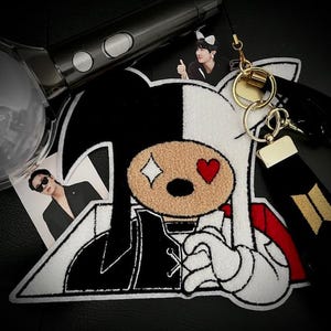 May include: Black and white embroidered patch of a cartoon character with a black and white face, one eye is a diamond and the other is a red heart. The character is wearing a black and white jacket with a white collar and a red scarf. The patch is on a black background.