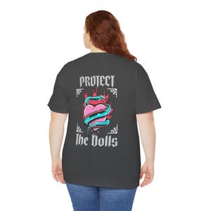 Puede incluir: Una camiseta gris oscuro jaspeada con un estampado gr&aacute;fico de un coraz&oacute;n con una serpiente enroscada a su alrededor. El coraz&oacute;n est&aacute; en llamas y el texto "Protect the Dolls" est&aacute; impreso debajo del coraz&oacute;n.
