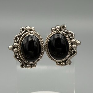 May include: A pair of oval-shaped earrings featuring a smooth, polished black stone set in a silver-toned metal frame. The setting is decorated with a twisted rope border and small spherical metal beads arranged along the outer edges.