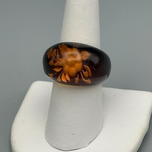 May include: A dark brown oval ring with an orange floral design. The ring is set on a white display stand, showcasing the intricate details of the carved flower pattern. The ring's glossy finish reflects light, highlighting the contrast between the dark base and the orange floral design.