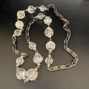 May include: A vintage necklace featuring clear, faceted glass beads of varying sizes, interspersed with silver-tone metal accents and a decorative chain. The necklace is displayed against a dark background, highlighting its intricate design and reflective qualities.
