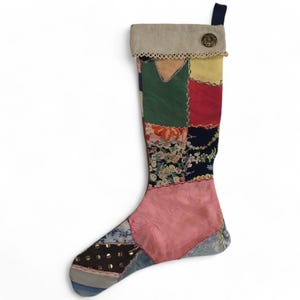 May include: A patchwork Christmas stocking with a variety of fabric patterns and colors, including pink, green, red, and floral prints. The top is a neutral beige with a decorative trim and a button. A navy blue loop is attached for hanging.
