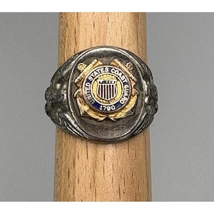 May include: A silver-toned ring featuring the United States Coast Guard emblem. The emblem includes the words "United States Coast Guard" and "1790" in a circular design with blue, gold, and red accents. The ring is displayed on a wooden dowel.