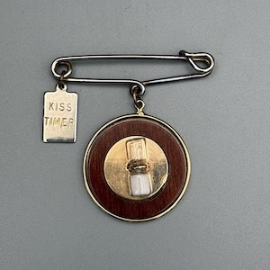 May include: A vintage brooch featuring a circular wooden pendant with a gold-colored center and a small vial. A rectangular tag with the words "KISS TIMER" hangs from the safety pin clasp. The brooch is a unique accessory.