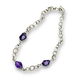 May include: A silver necklace featuring three faceted, deep purple amethyst gemstones. The chain alternates between simple and twisted links, with a clasp closure. The necklace is displayed against a white background.