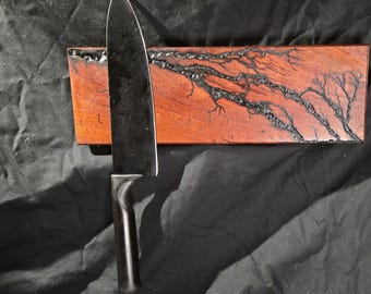 May include: A black knife with a wooden handle is attached to a magnetic knife holder. The holder is made of dark wood with black lightning-like patterns.