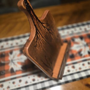 May include: A handcrafted wooden cutting board with a handle. The board features a unique tree-like design burned into the wood. The board is brown and has a natural finish. The cutting board is displayed on a decorative table runner.