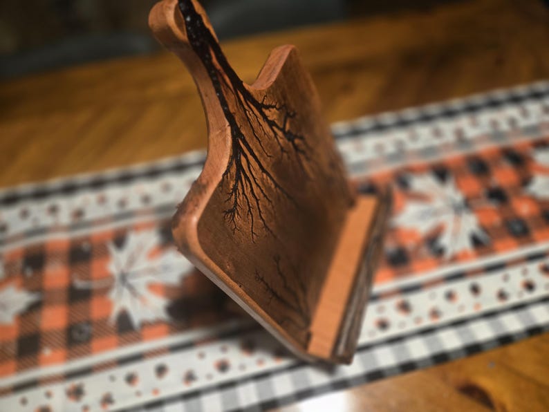 May include: A handcrafted wooden cutting board with a handle. The board features a unique tree-like design burned into the wood. The board is brown and has a natural finish. The cutting board is displayed on a decorative table runner.