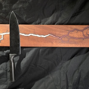 May include: A wooden magnetic knife holder with a white and red resin inlay. The holder has a natural wood finish and a sleek, modern design. A black kitchen knife is attached to the holder.